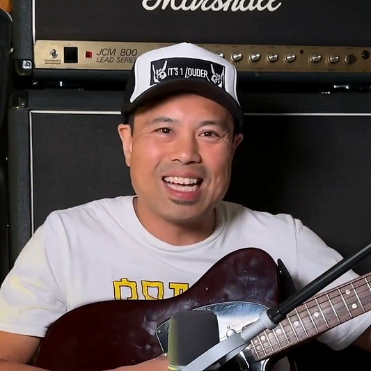 PJ Pat smiling with a guitar in front of a Marshall stack, wearing the It's 1 Louder B/W Trucker Hat with white front and black rock horns patch