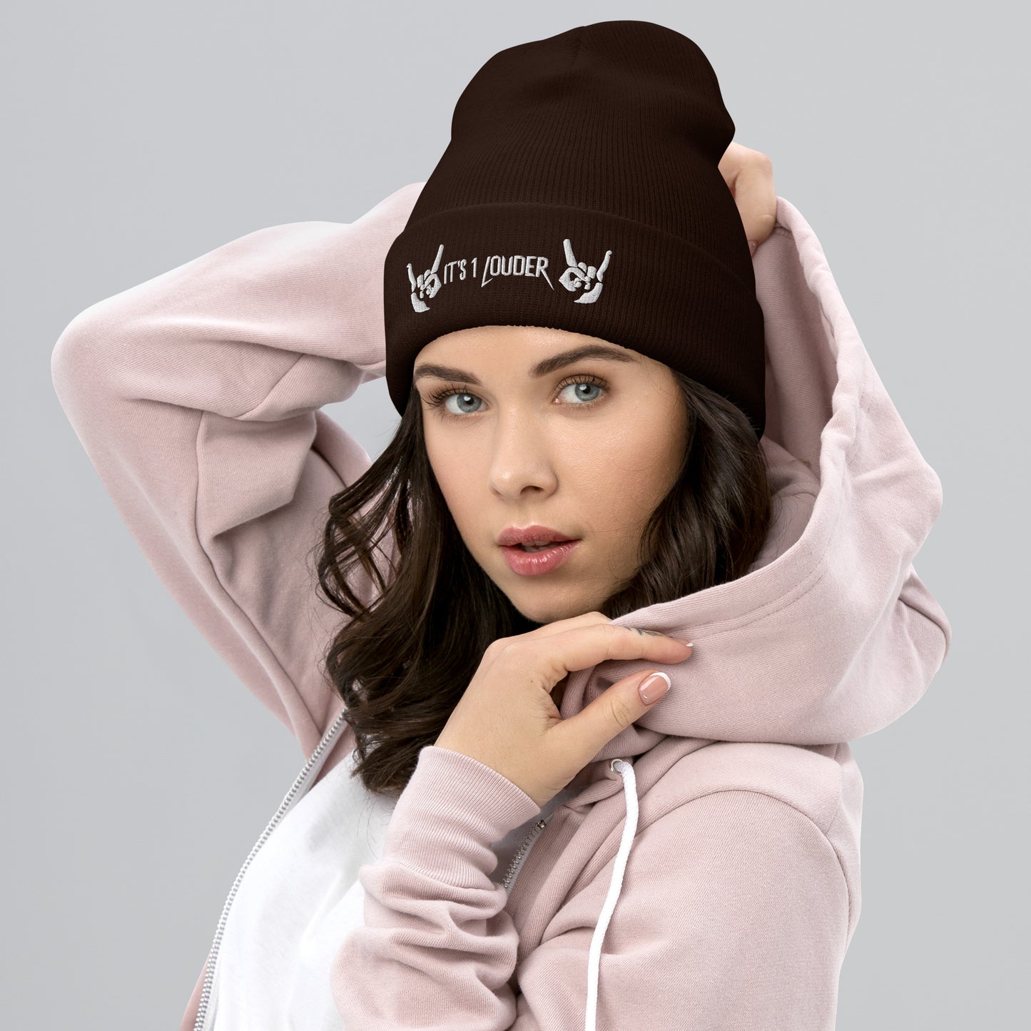 Caucasian woman looking straight at the viewer wearing a maroon beanie. There are also two hands embroidered onto the beanie showing the rock and roll symbol. In between those hands are the words embroidered saying "It's 1 Louder". This beanie gives her a rock n roll attire or a punk look.